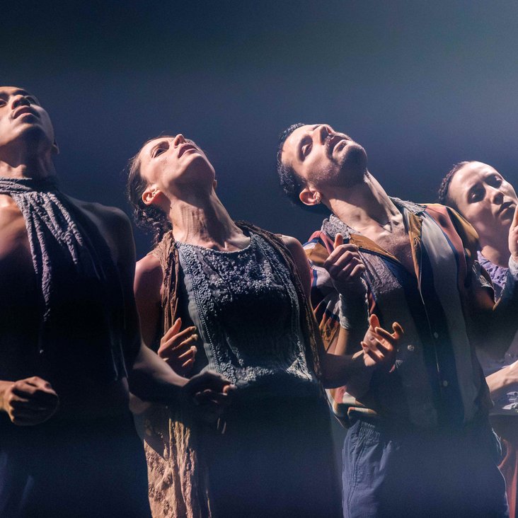Die Tänzer:innen von Gauthier Dance in verschiedenfarbigen Kostümen auf der Bühne. Die Bühne ist auch dunkel und die Beleuchtung ist atmosphärisch.