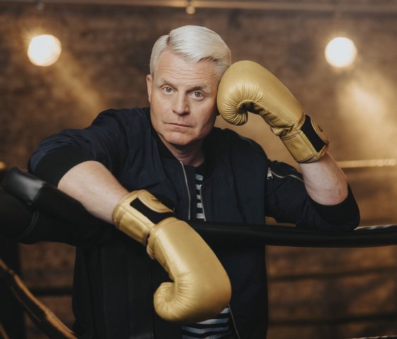 Der blonde Entertainer Guido Cantz lehnt sich über die Abtrennung eines Boxrings. Er trägt goldene Handschuhe. Er wirkt abgeschafft.