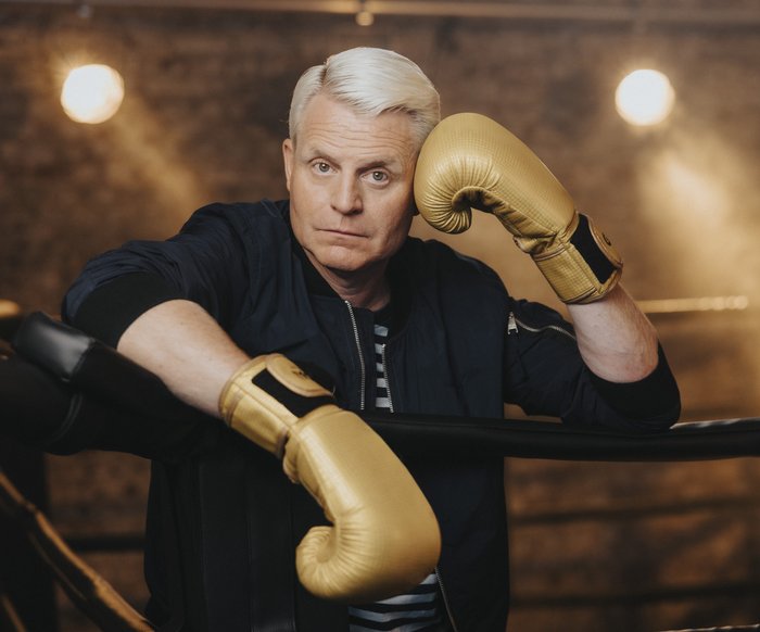 Der blonde Entertainer Guido Cantz lehnt sich über die Abtrennung eines Boxrings. Er trägt goldene Handschuhe. Er wirkt abgeschafft.