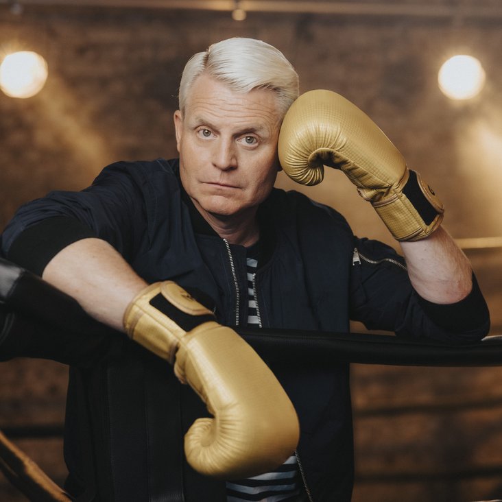 Der blonde Entertainer Guido Cantz lehnt sich über die Abtrennung eines Boxrings. Er trägt goldene Handschuhe. Er wirkt abgeschafft.