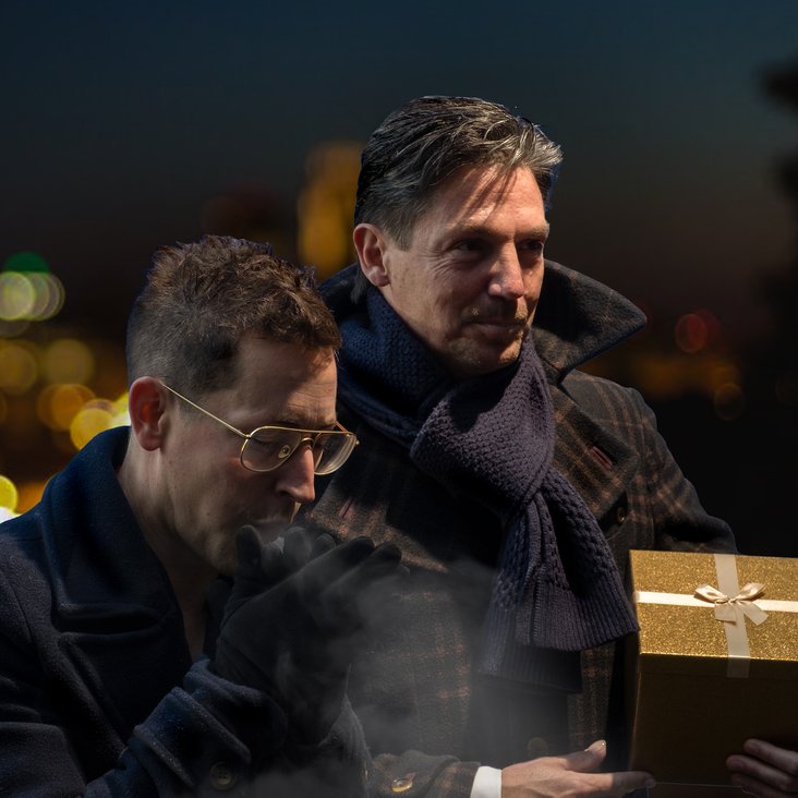 Die beiden Café del Mundo Musiker stehen in Wintermänteln und mit einem Geschenk draußen vor einer Lichterkulisse