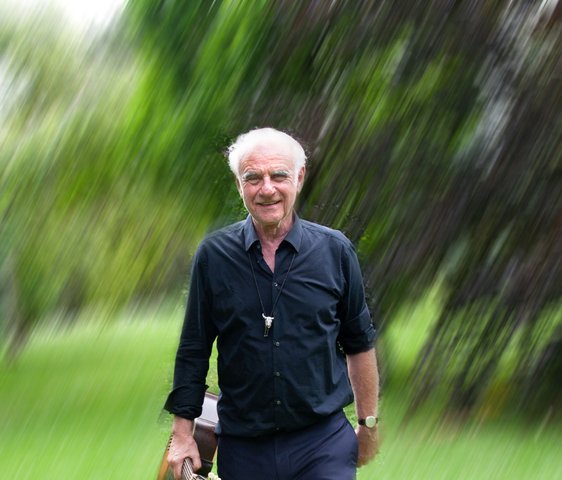 Roland Baisch in dunkler Kleidung läuft über eine grüne Wiese, im Hintergrund sind Bäume zu sehen. Er hat eine Gitarre in der Hand. Das ganze Bild ist etwas unscharf gestaltet.