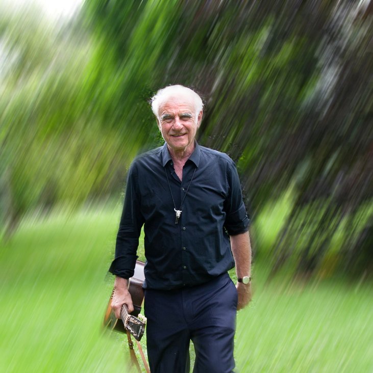 Roland Baisch in dunkler Kleidung läuft über eine grüne Wiese, im Hintergrund sind Bäume zu sehen. Er hat eine Gitarre in der Hand. Das ganze Bild ist etwas unscharf gestaltet.