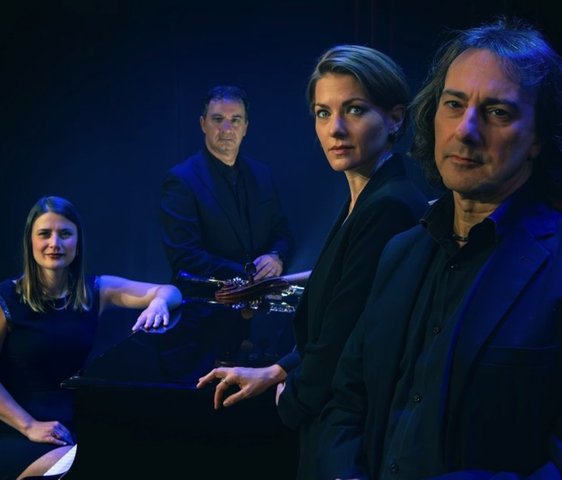 Das Quartett schaut in die Kamera, der Hintergrund ist schwarz und die Beleuchtung blau.