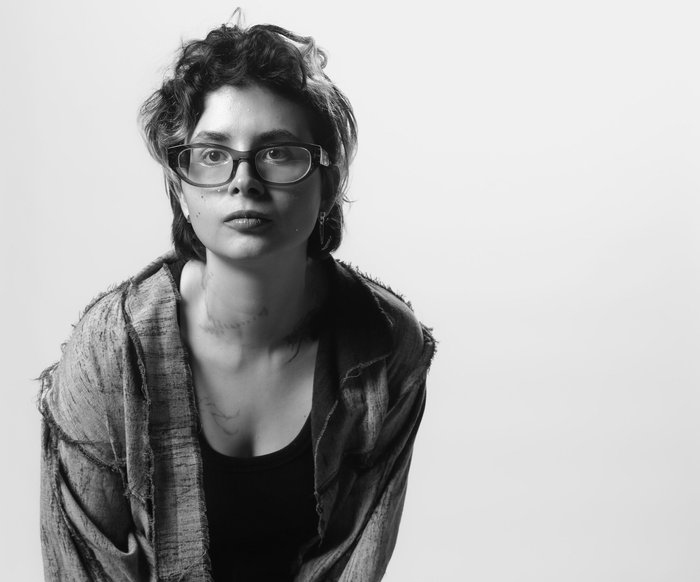 Schwarzweißporträt einer Person, sie trägt ein offenes Hemd über einem dunklen Top und eine Jogginghose mit Streifen an der Seite. Stehende Pose vor neutralem Studiohintergrund.