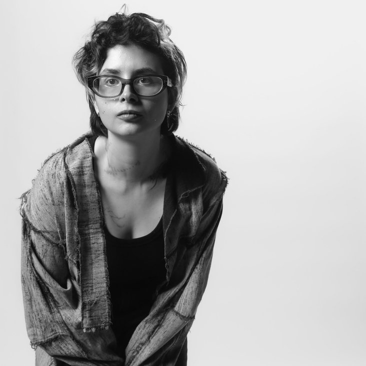 Schwarzweißporträt einer Person, sie trägt ein offenes Hemd über einem dunklen Top und eine Jogginghose mit Streifen an der Seite. Stehende Pose vor neutralem Studiohintergrund.