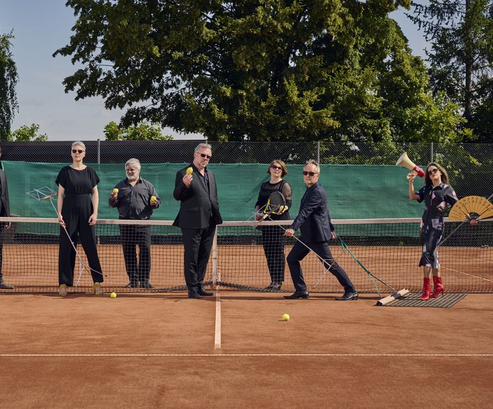 Die Künstler stehen auf einem Tennisplatz am Netz. Drei stehen hinter dem Netzt und vier davor. Sie sind alle in schwarz gekleidet und tragen Sonnenbrillen.