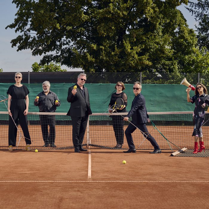 Die Künstler stehen auf einem Tennisplatz am Netz. Drei stehen hinter dem Netzt und vier davor. Sie sind alle in schwarz gekleidet und tragen Sonnenbrillen.