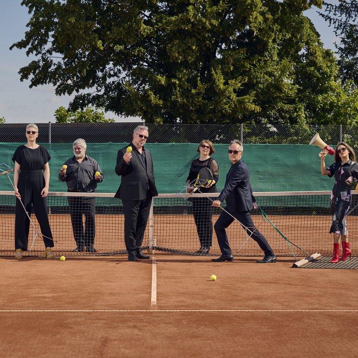 Die Künstler stehen auf einem Tennisplatz am Netz. Drei stehen hinter dem Netzt und vier davor. Sie sind alle in schwarz gekleidet und tragen Sonnenbrillen.