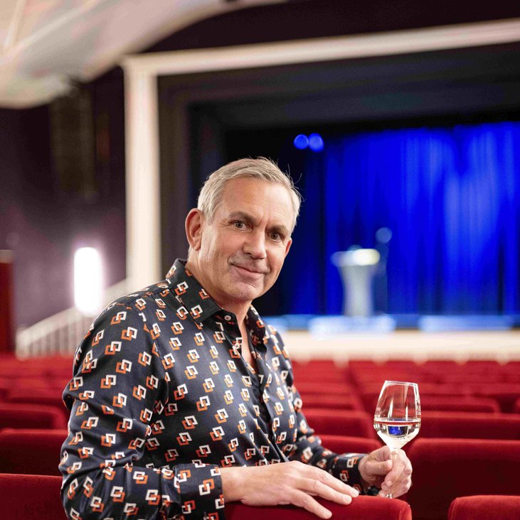 Wladimir Kaminer steht mit einem Weinglas in der Hand im Zuschauerraum eines Theaters