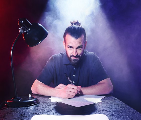 David Kebekus sitzt an einem Tisch, vor ihm Stift und Papier, neben ihm eine Schreibtisch-Lampe. Er blickt in die Kamera.