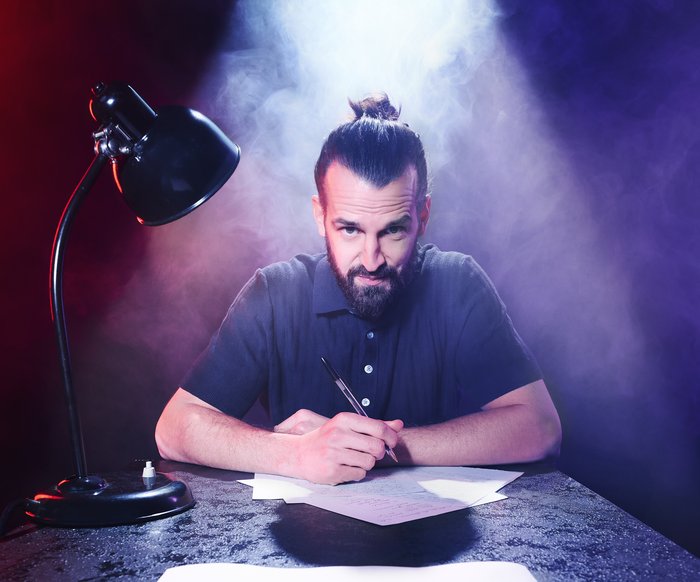 David Kebekus sitzt an einem Tisch, vor ihm Stift und Papier, neben ihm eine Schreibtisch-Lampe. Er blickt in die Kamera.