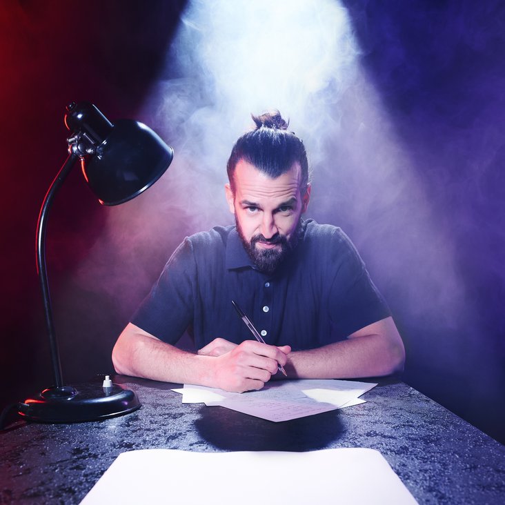 David Kebekus sitzt an einem Tisch, vor ihm Stift und Papier, neben ihm eine Schreibtisch-Lampe. Er blickt in die Kamera.