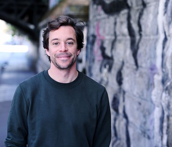 Tobias Krell steht im urbanen Raum neben einer Wand, auf der ein Graffiti zu erkennen ist. Er lächelt in die Kamera.