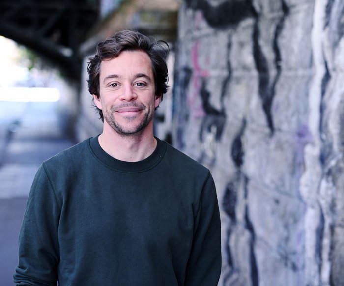 Tobias Krell steht im urbanen Raum neben einer Wand, auf der ein Graffiti zu erkennen ist. Er lächelt in die Kamera.