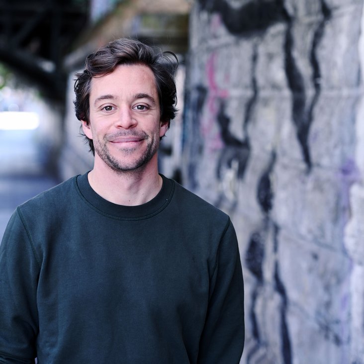 Tobias Krell steht im urbanen Raum neben einer Wand, auf der ein Graffiti zu erkennen ist. Er lächelt in die Kamera.