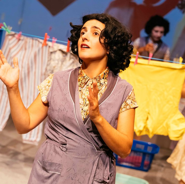 Frau gestikuliert mit erhobenen Händen auf der Bühne, andere Frauen im Hintergrund zwischen Wäscheleinen