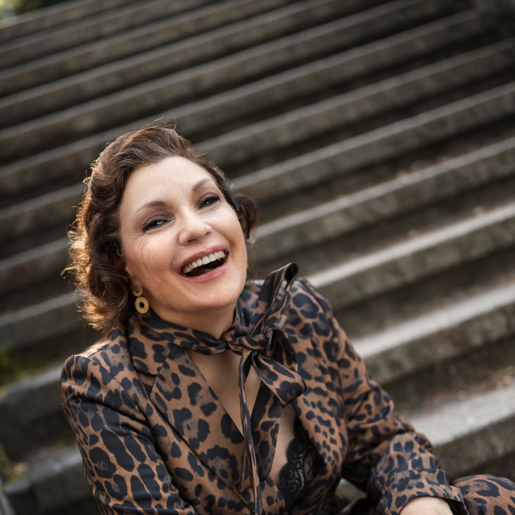Portraitfoto Claudia Carbo, sie sitzt auf Treppenstufen und trägt Hose und Bluse im Leopardenmuster. Sie lacht in die Kamera.