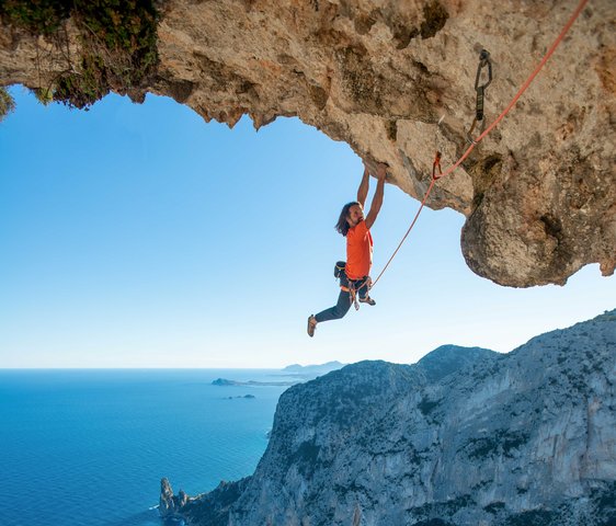 Alexander Huber hält sich mit beiden Händen an einem großen Felsvorsprung fest, im Hintergrund Felsküste und das Meer. Es ist mit einem Seil gesichert.