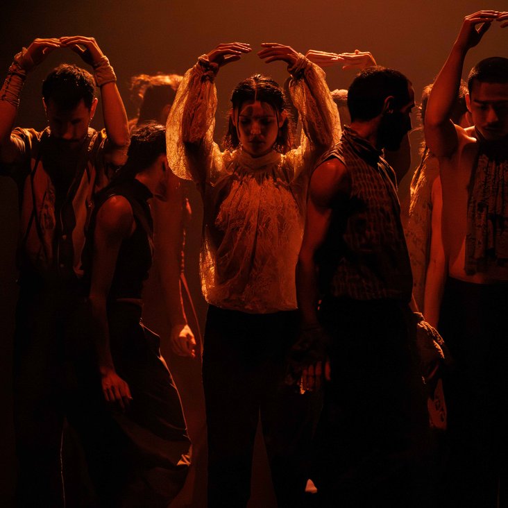Die Tänzer:innen von Gauthier Dance in verschiedenfarbigen Kostümen auf der Bühne. Die Bühne ist auch dunkel und die Beleuchtung ist atmosphärisch.