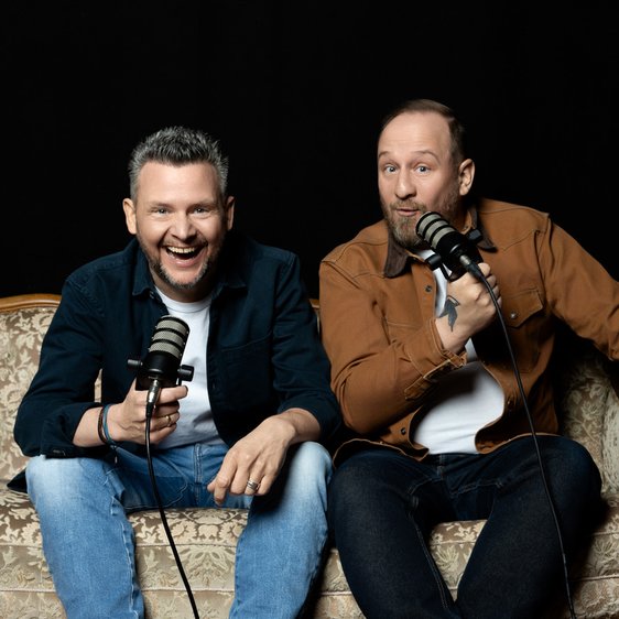 Philip Simon und Tobias Mann sitzen auf einem Sofa und halten Mikros in der Hand, der Hintergrund ist dunkel.