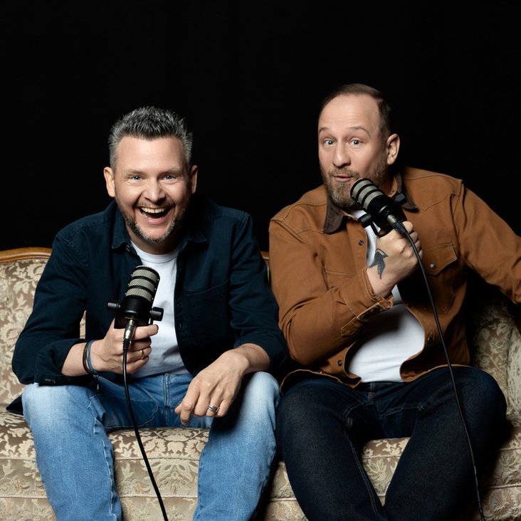 Philip Simon und Tobias Mann sitzen auf einem Sofa und halten Mikros in der Hand, der Hintergrund ist dunkel.