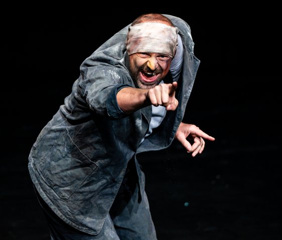 Der Künstler schaut leicht vorgebeugt in die Kamera, während er mit dem rechten Arm geradeaus zeigt.