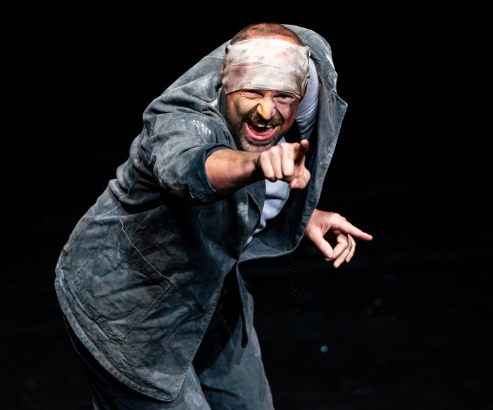 Der Künstler schaut leicht vorgebeugt in die Kamera, während er mit dem rechten Arm geradeaus zeigt.