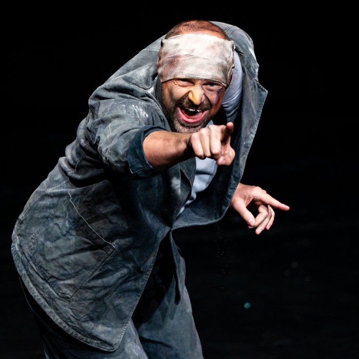 Der Künstler schaut leicht vorgebeugt in die Kamera, während er mit dem rechten Arm geradeaus zeigt.