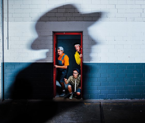 Drei Männer, verkleidet als Jungen, blicken durch eine Tür. Auf der Wand der Türe befindet sich ein geheimnisvoller Schatten eines Mannes.