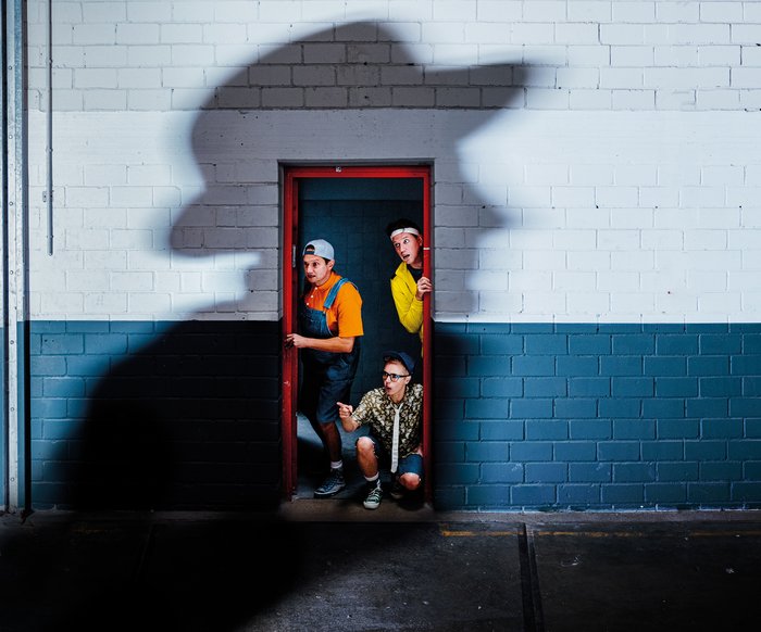 Drei Männer, verkleidet als Jungen, blicken durch eine Tür. Auf der Wand der Türe befindet sich ein geheimnisvoller Schatten eines Mannes.