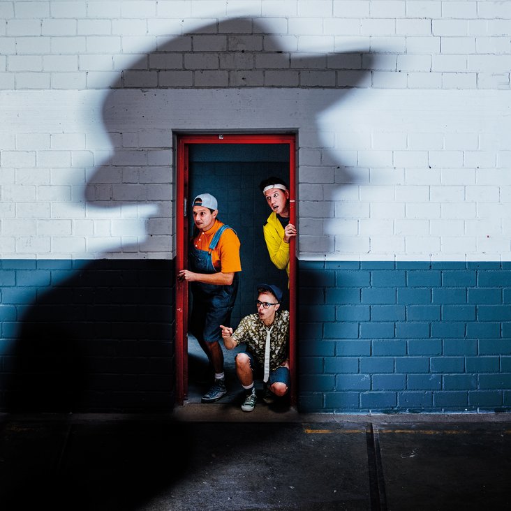 Drei Männer, verkleidet als Jungen, blicken durch eine Tür. Auf der Wand der Türe befindet sich ein geheimnisvoller Schatten eines Mannes.