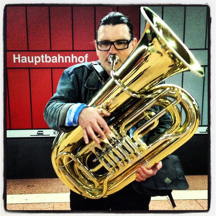 Ein Mann spielt Tuba. Er steht an einem Gleis. Im Hintergrund ist der Schriftzug "Hauptbahnhof" an der Wand zu lesen.