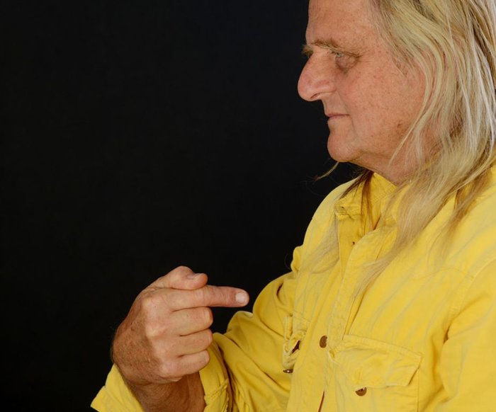 Portraitfoto von Büdi Siebert. Man sieht ihn im seitlichen Profil, er trägt ein gelber Hemd und zeigt mit dem Zeigefinger auf sich selbst.