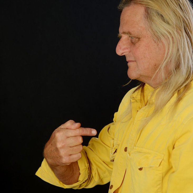 Portraitfoto von Büdi Siebert. Man sieht ihn im seitlichen Profil, er trägt ein gelber Hemd und zeigt mit dem Zeigefinger auf sich selbst.