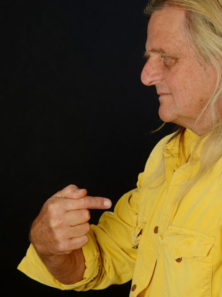 Portraitfoto von Büdi Siebert. Man sieht ihn im seitlichen Profil, er trägt ein gelber Hemd und zeigt mit dem Zeigefinger auf sich selbst.
