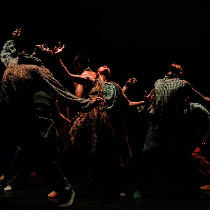 Die Tänzer:innen von Gauthier Dance in verschiedenfarbigen Kostümen auf der Bühne. Die Bühne ist auch dunkel und die Beleuchtung ist atmosphärisch.