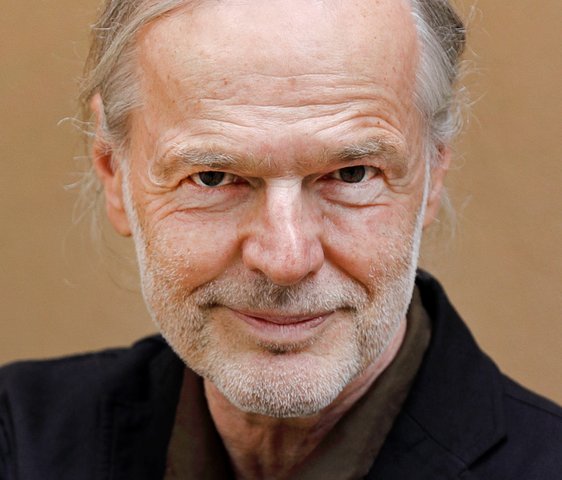 Ein Portrait eines Mannes. Er hat einen grauen Stoppelbart und graue lange Haare. Er trägt ein schwarzes Hemd und lächelt verschmitzt.