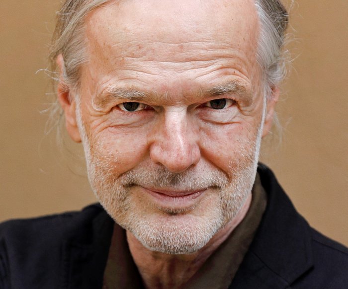 Ein Portrait eines Mannes. Er hat einen grauen Stoppelbart und graue lange Haare. Er trägt ein schwarzes Hemd und lächelt verschmitzt.