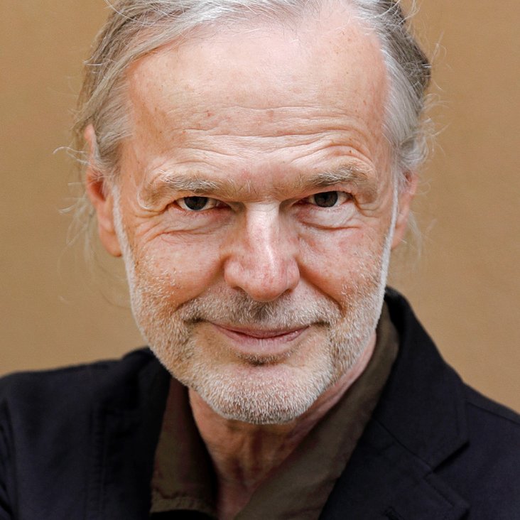Ein Portrait eines Mannes. Er hat einen grauen Stoppelbart und graue lange Haare. Er trägt ein schwarzes Hemd und lächelt verschmitzt.