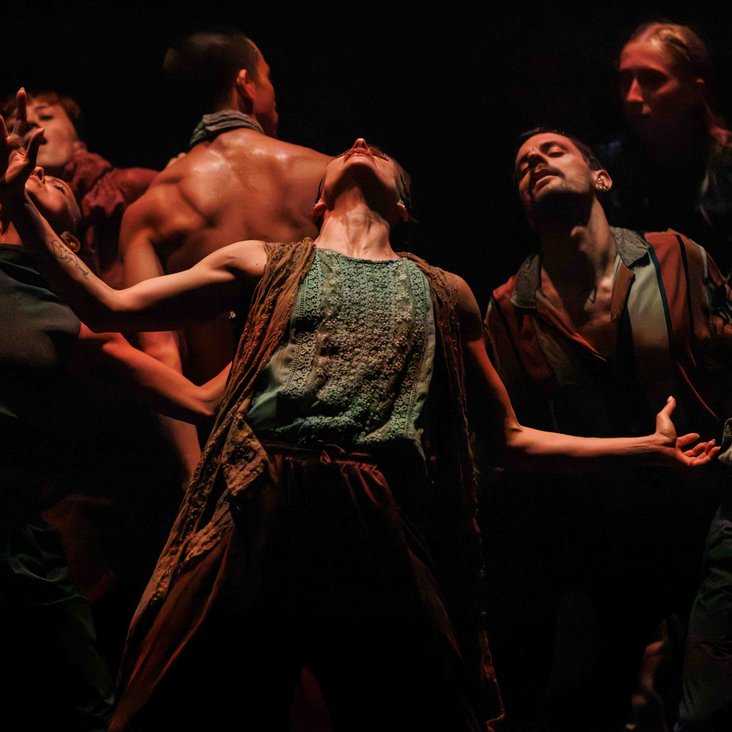 Die Tänzer:innen von Gauthier Dance in verschiedenfarbigen Kostümen auf der Bühne. Die Bühne ist auch dunkel und die Beleuchtung ist atmosphärisch.