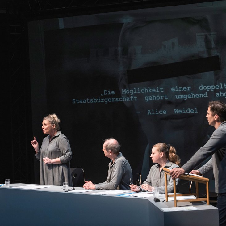 Szenenfoto, es zeigt fünf Schauspieler:innen sitzend und stehend in grauer Kleidung auf der Bühne. Im Hintergrund das schwarz-weiß Foto einer Person, die Augen sind zensiert.