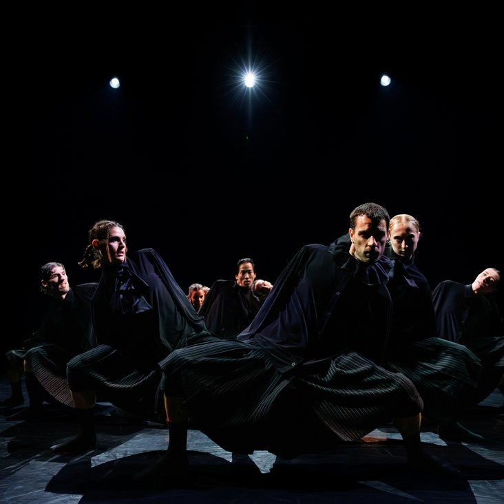 Die Tänzer:innen von Gauthier Dance in schwarzen Kostümen auf der Bühne. Die Bühne ist auch dunkel und es gibt wenig Licht.
