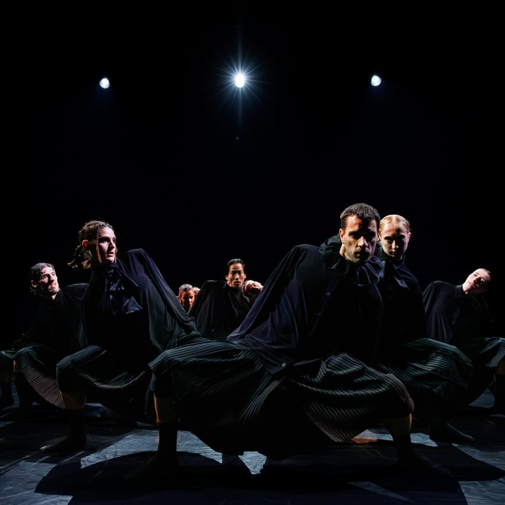 Die Tänzer:innen von Gauthier Dance in schwarzen Kostümen auf der Bühne. Die Bühne ist auch dunkel und es gibt wenig Licht.