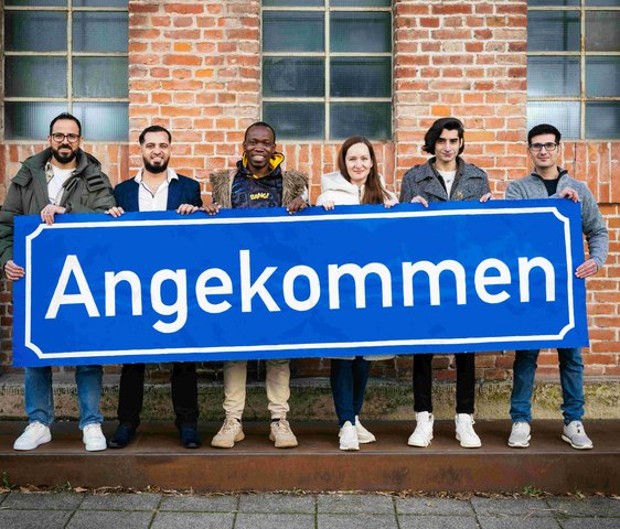 Die Teilnehmen der Produktion "Angekommen" stehen vor der Backsteinwand des Theaterhauses, sie halten alle gemeinsam ein großes blaues Schild mit der Aufschrift "Angekommen" in den Händen