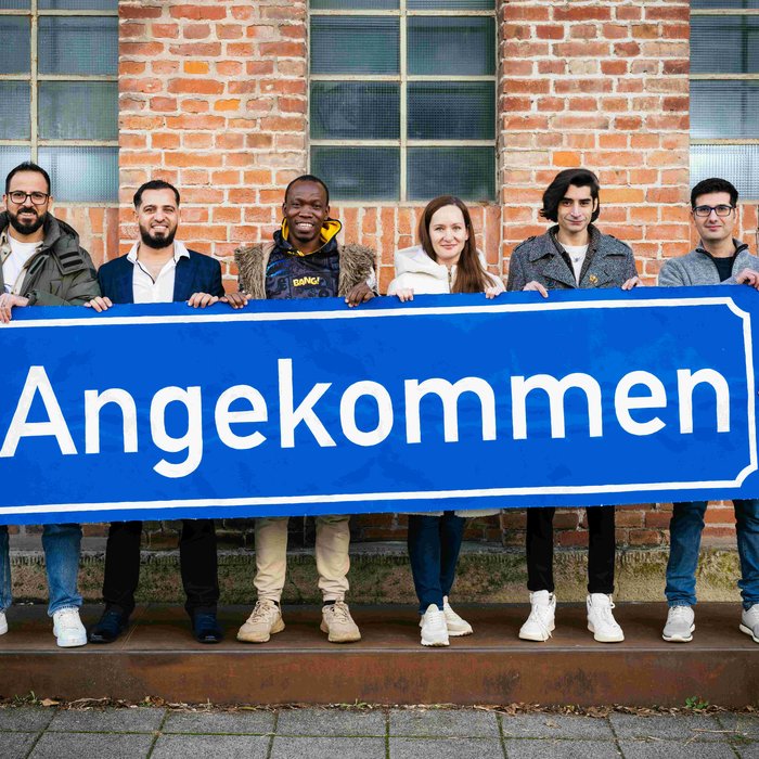 Die Teilnehmen der Produktion "Angekommen" stehen vor der Backsteinwand des Theaterhauses, sie halten alle gemeinsam ein großes blaues Schild mit der Aufschrift "Angekommen" in den Händen