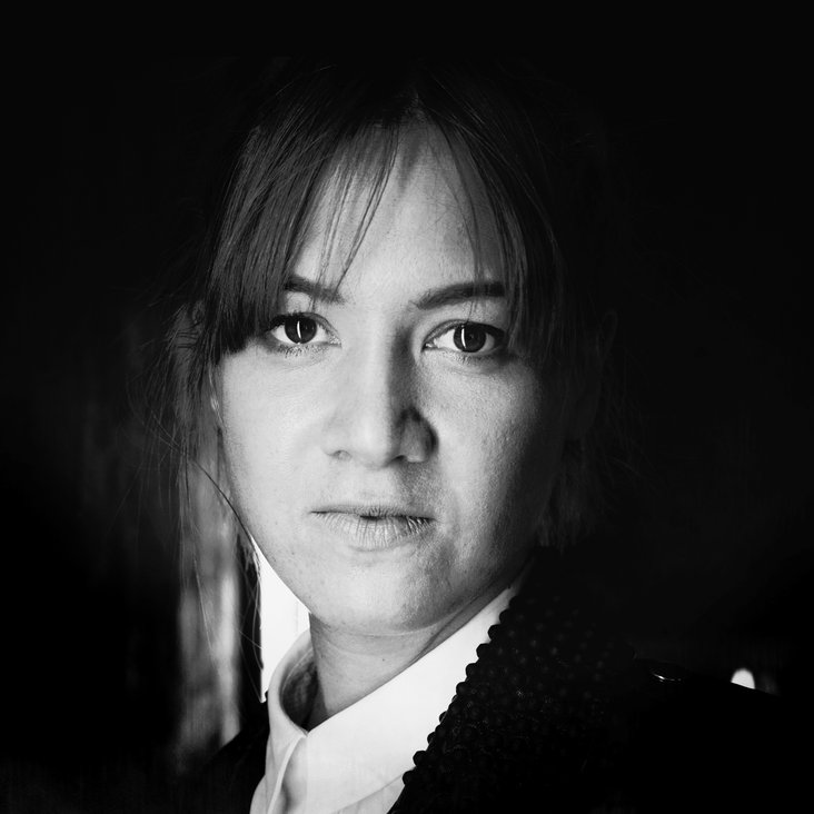Ein schwarz weiß Portrait einer Frau. Sie trägt eine weiße Bluse, Strähnen eines leichten Ponys fallen ihr ins Gesicht.