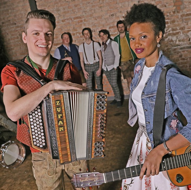 Zwei Musiker:innen mit Akkordeon und Gitarre stehen im Vordergrund, im Hintergrund sind vier weitere Musiker zu sehen