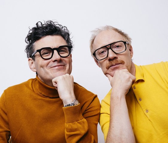 Manuel Ruby und Simon Schwarz, sie tragen einen braunen Pullover bzw. ein gelbes Hemd und blicken in die Kamera.