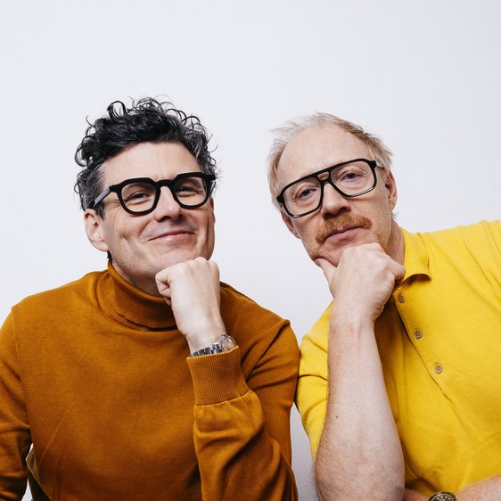 Manuel Ruby und Simon Schwarz, sie tragen einen braunen Pullover bzw. ein gelbes Hemd und blicken in die Kamera.